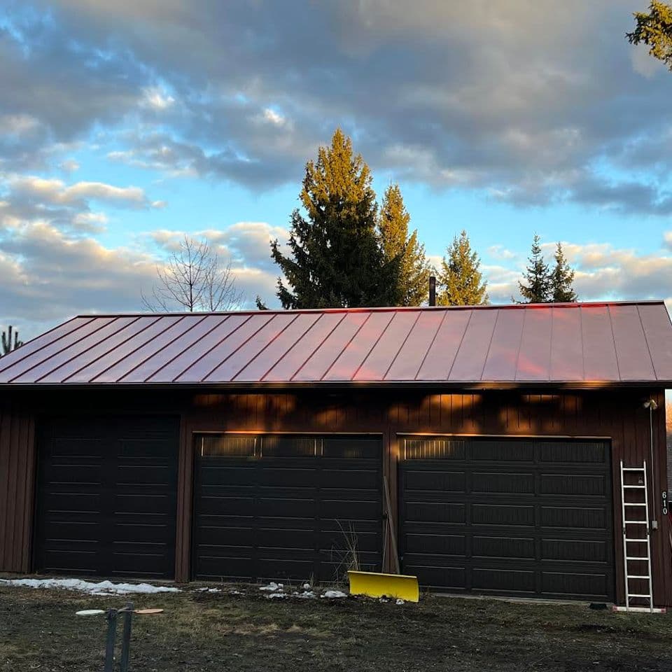 Modern three-car garage with a metal roof under a cloudy sky and pine trees.