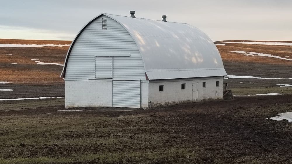 White barn on a rural landscape with snow patches and golden grass under a sunset sky.
