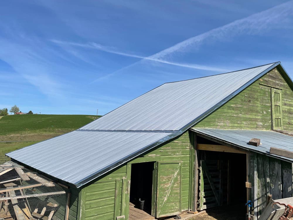 Green barn with a metal roof under a blue sky and wispy clouds. Countryside landscape visible.