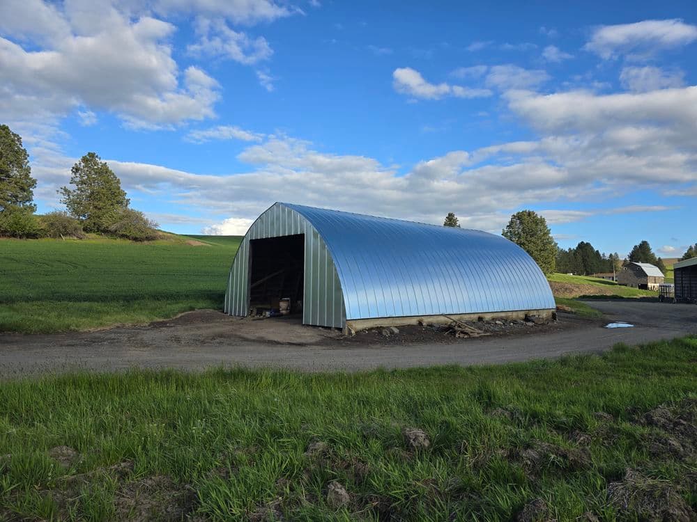 Metal barn with curved roof on a gravel path, surrounded by green fields and trees.