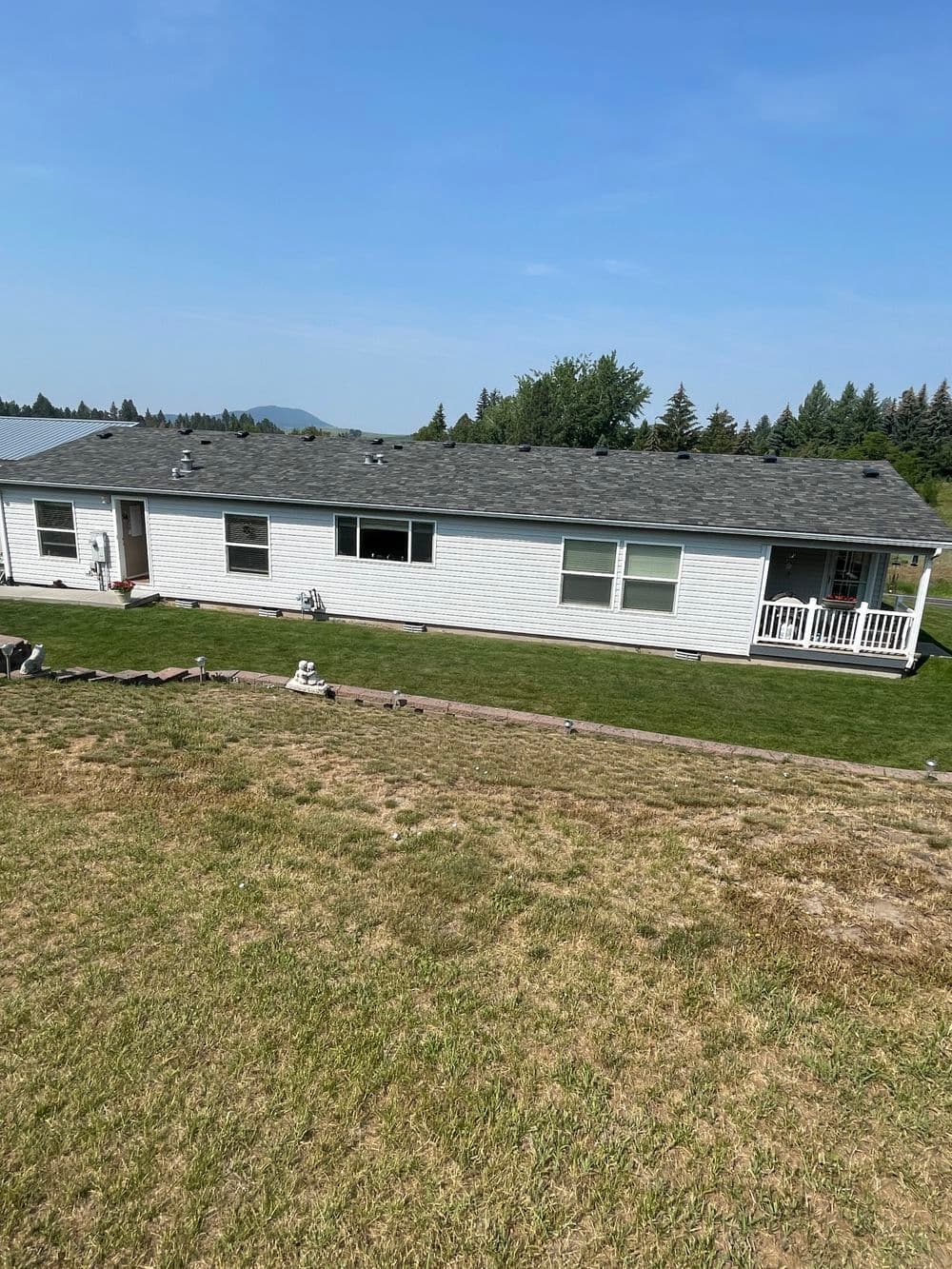 Single-story white mobile home with a porch, surrounded by green grass and trees in the background.