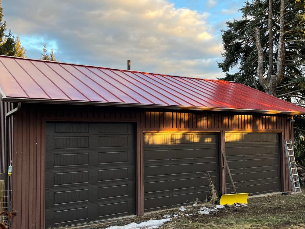 Red metal roof garage with three dark doors and ladder, set against a scenic sky.