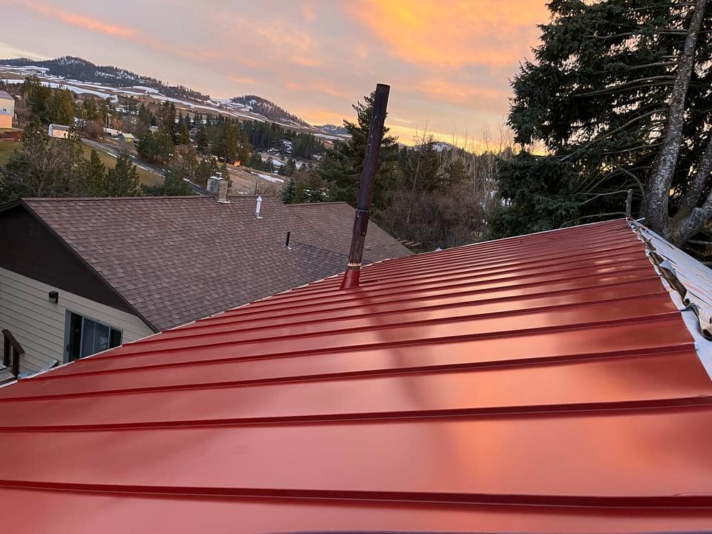 Red metal roof with chimney at sunset, overlooking a scenic landscape and distant hills.